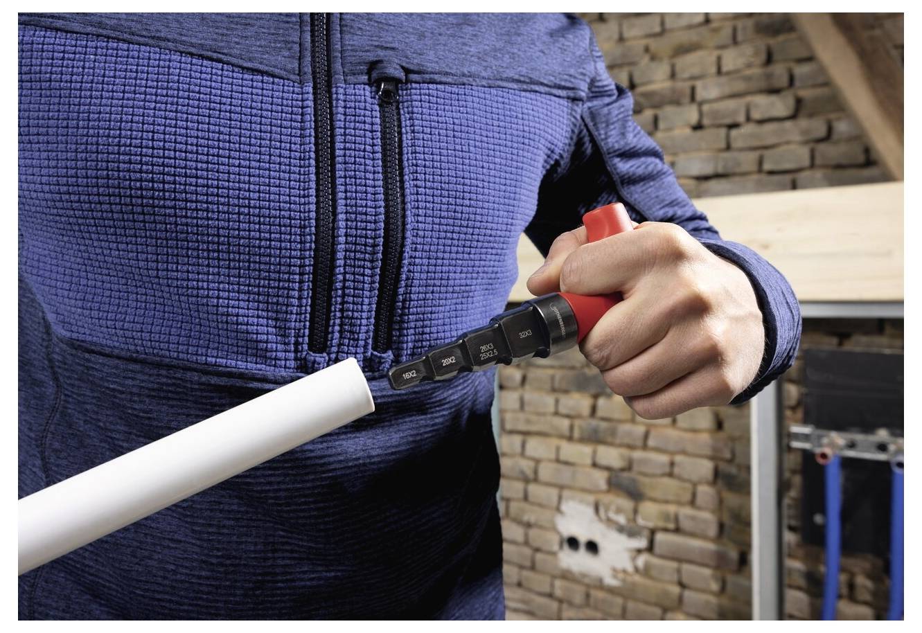 Eine Person in einer blauen Jacke verwendet ein Werkzeug, um ein weißes Rohr drinnen in der Nähe einer Backsteinwand unter Renovierung zu schneiden.