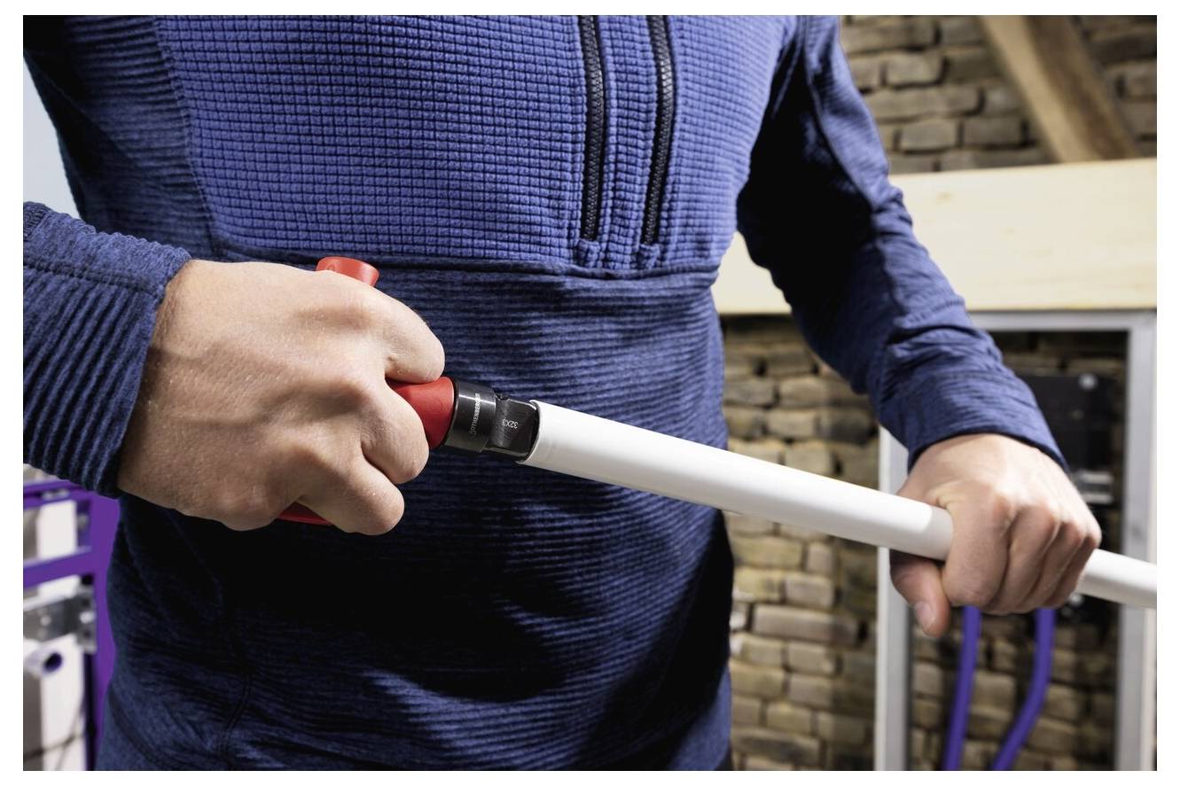 Eine Person in einem blauen Pullover schneidet ein weißes Rohr mit einem roten Rohrabschneider vor einem Hintergrund aus gestapelten Steinen und Holzbalken.
