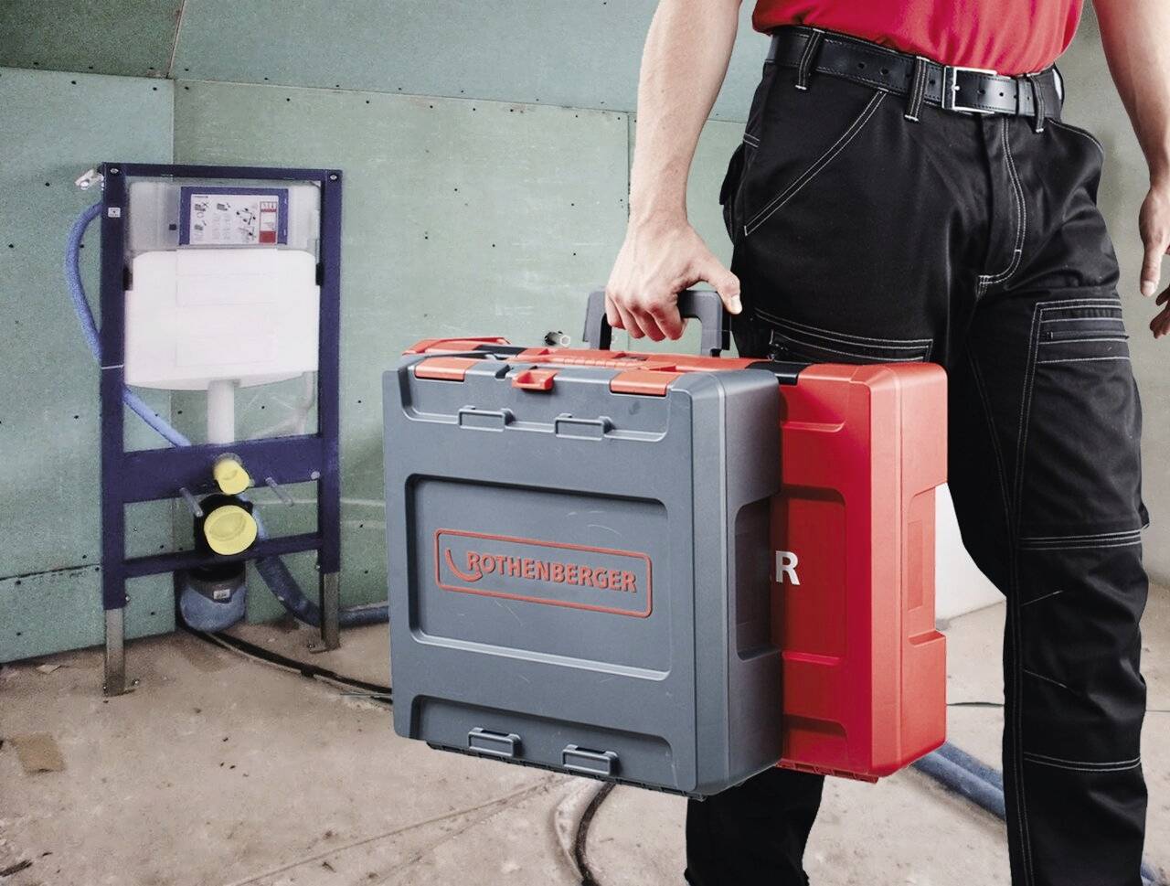 „Person in Arbeitskleidung, die eine rote und graue Rothenberger-Werkzeugkiste hält und in der Nähe von Sanitärinstallationsausrüstung in einer Baustellenumgebung steht.