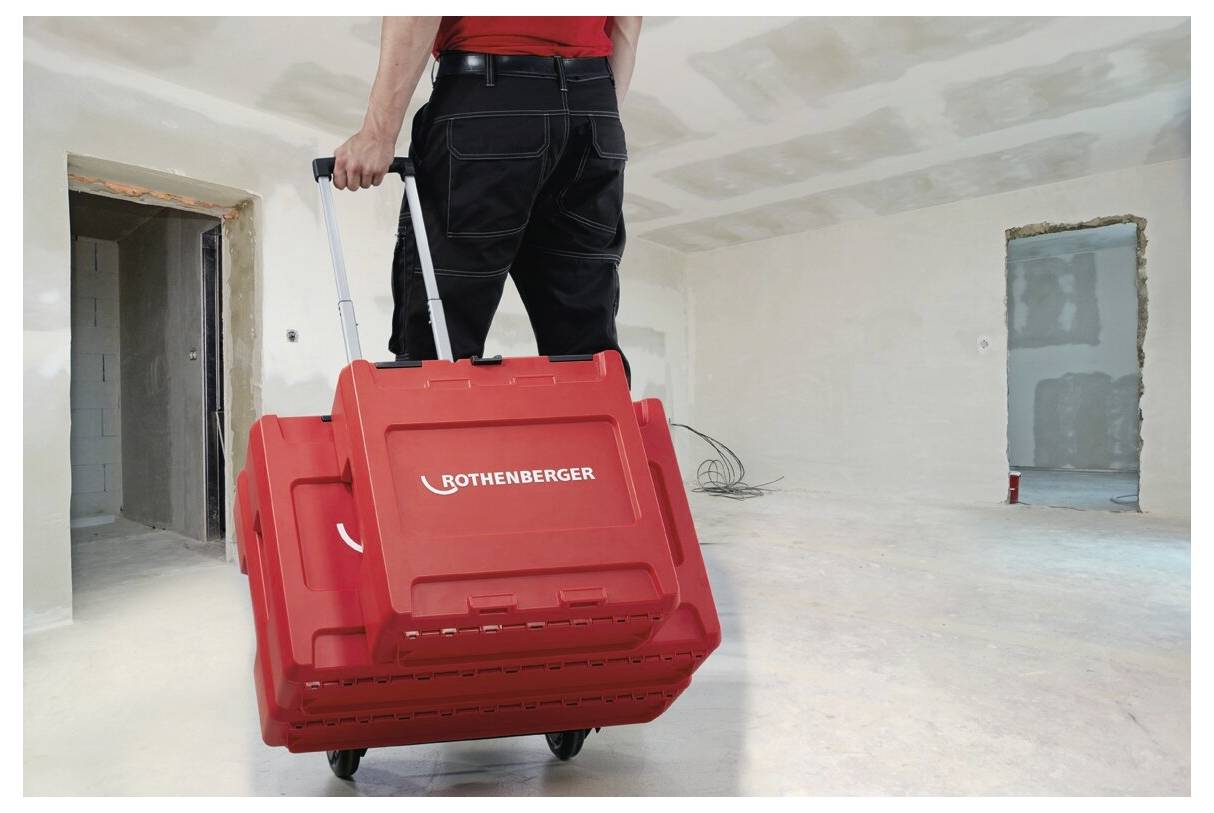 Eine Person zieht einen roten Werkzeugkoffer mit der Aufschrift 'Rothenberger' durch einen teilweise fertiggestellten Raum mit ungestrichenen Wänden und Baumaterialien.