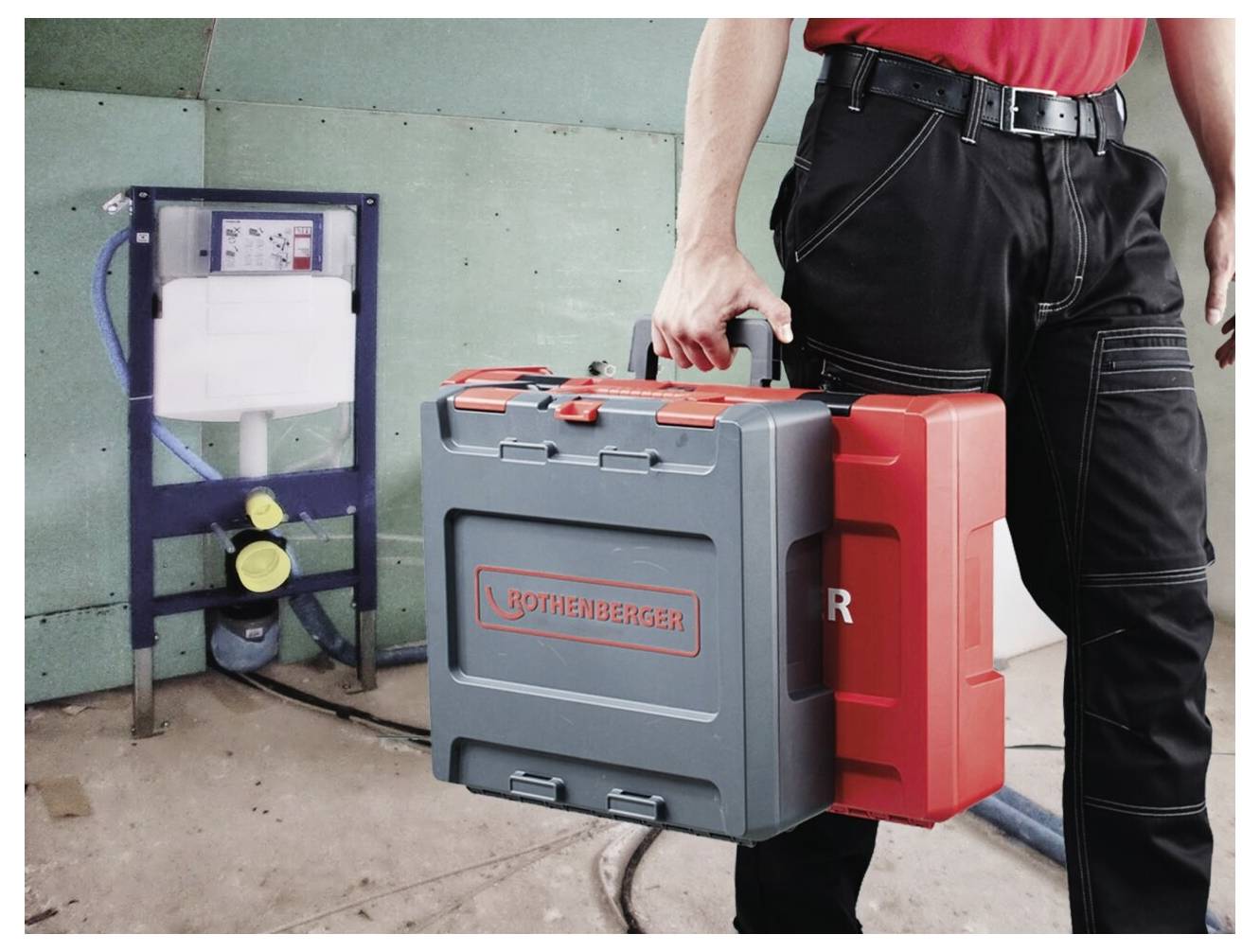 Eine Person in Arbeitskleidung trägt zwei Werkzeugkästen vor einer wandmontierten Sanitärinstallation, was auf eine Sanitärreparatur oder -installation hindeutet.