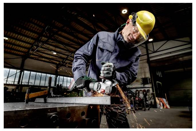 Ein Arbeiter mit Schutzhelm und Handschuhen schleift Metall, wobei Funken sprühen, in einem großen industriellen Lagergebäude.