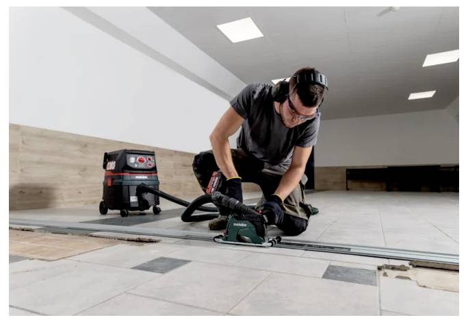 Eine Person mit Sicherheitsausrüstung verwendet ein Elektrowerkzeug, um Fliesen auf dem Boden zu schneiden, während ein Staubsauger in der Nähe steht und eine Bauaktivität anzeigt.