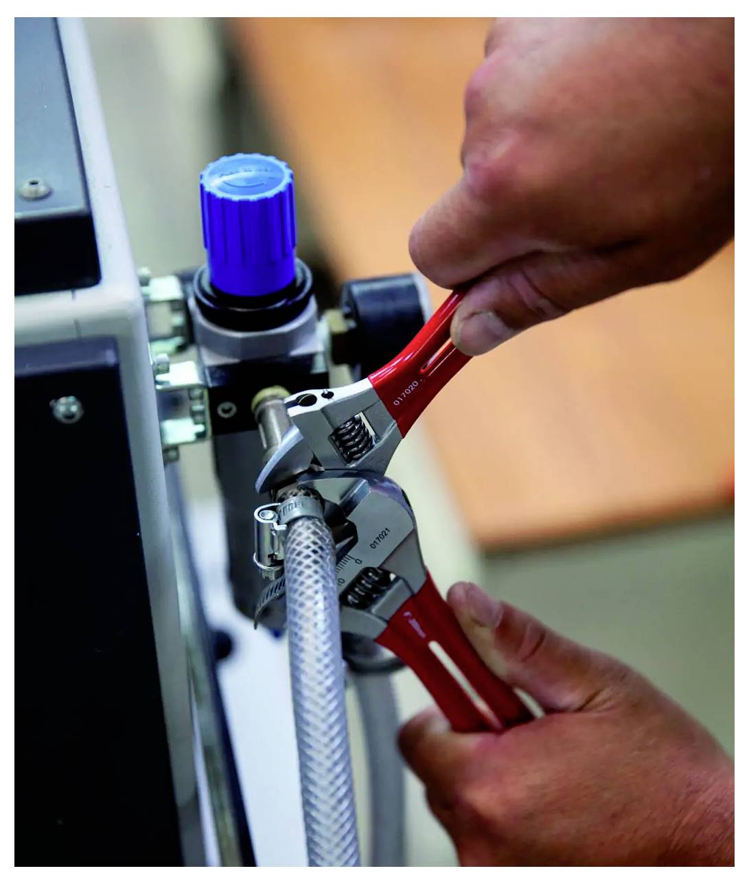 Hände mit roten verstellbaren Schraubenschlüsseln, die eine Schlauchverbindung an einer Industriemaschine anziehen.