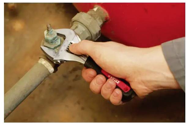 Hand, die mit einem Schraubenschlüssel eine Rohrverschraubung an einem Metallventil gegen einen roten Tank anzieht.