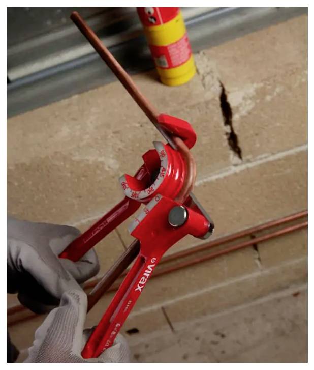 Behandschuhte Hand, die einen roten Rohrbender verwendet, um ein Kupferrohr gegen eine Ziegelmauer zu biegen.
