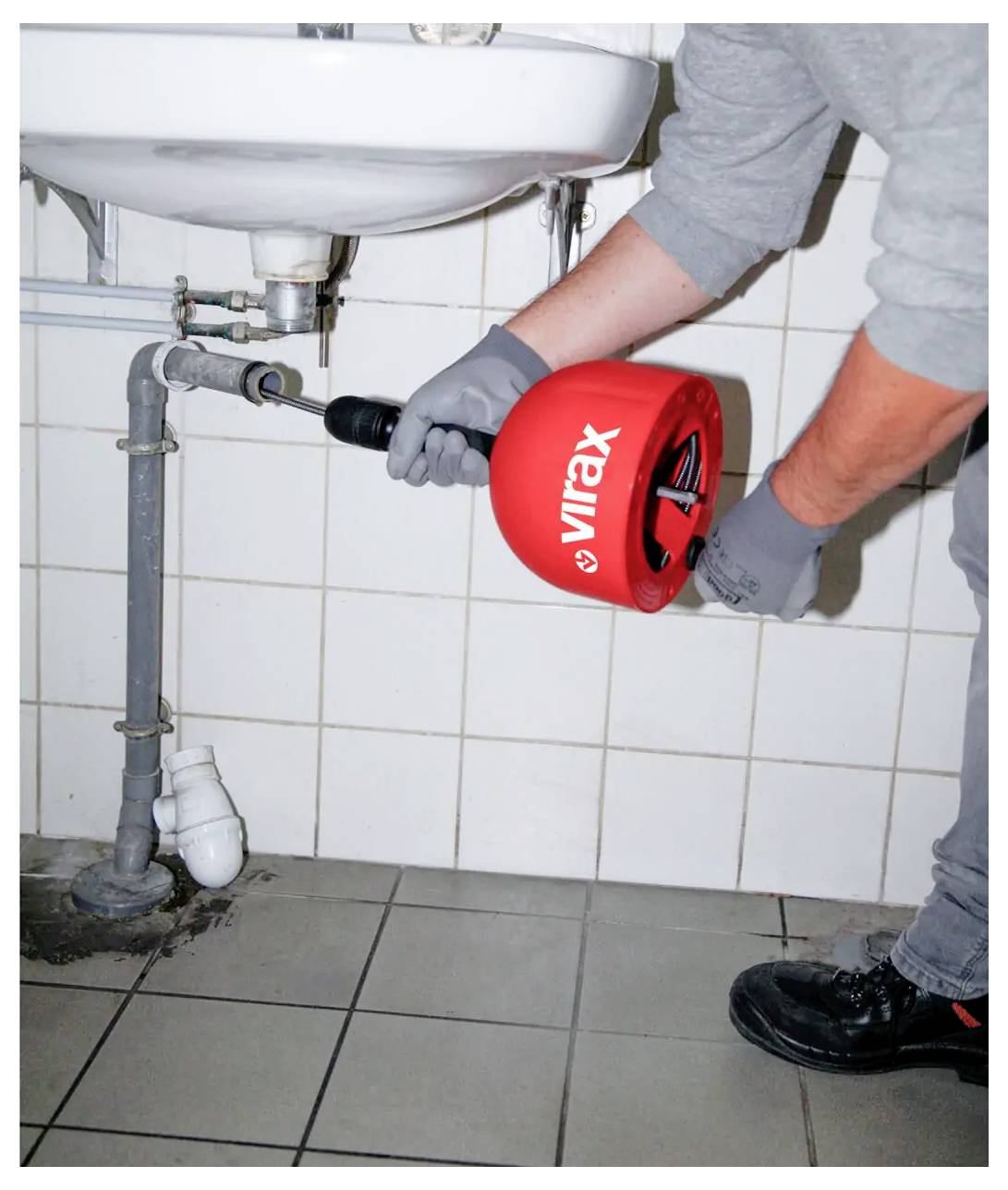 Klempner mit einer roten Virax-Rohrreinigungsspirale beim Freimachen eines verstopften Badezimmer-Waschbeckens unter einem wandmontierten Becken.