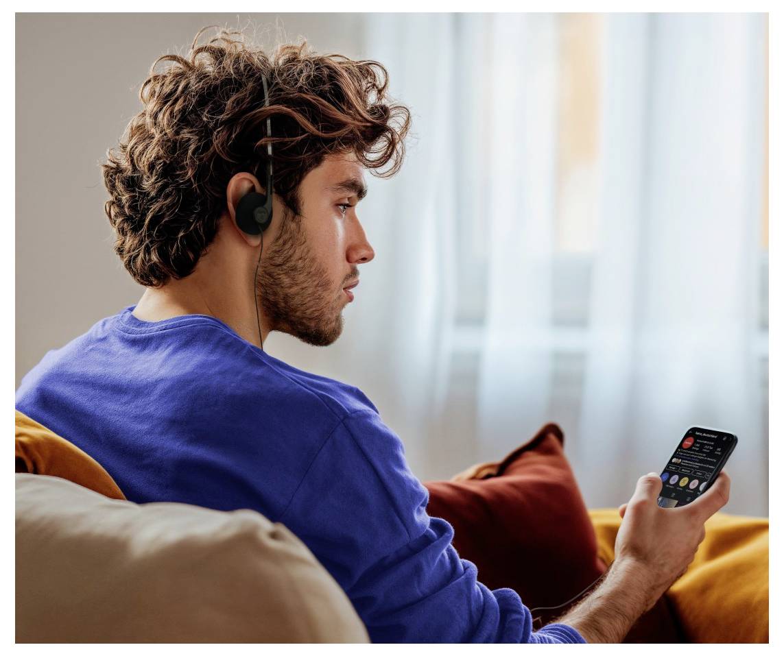 Eine Person mit Kopfhörern sitzt auf einem Sofa und blickt auf einen Smartphone-Bildschirm in einem gemütlichen, hell erleuchteten Raum.