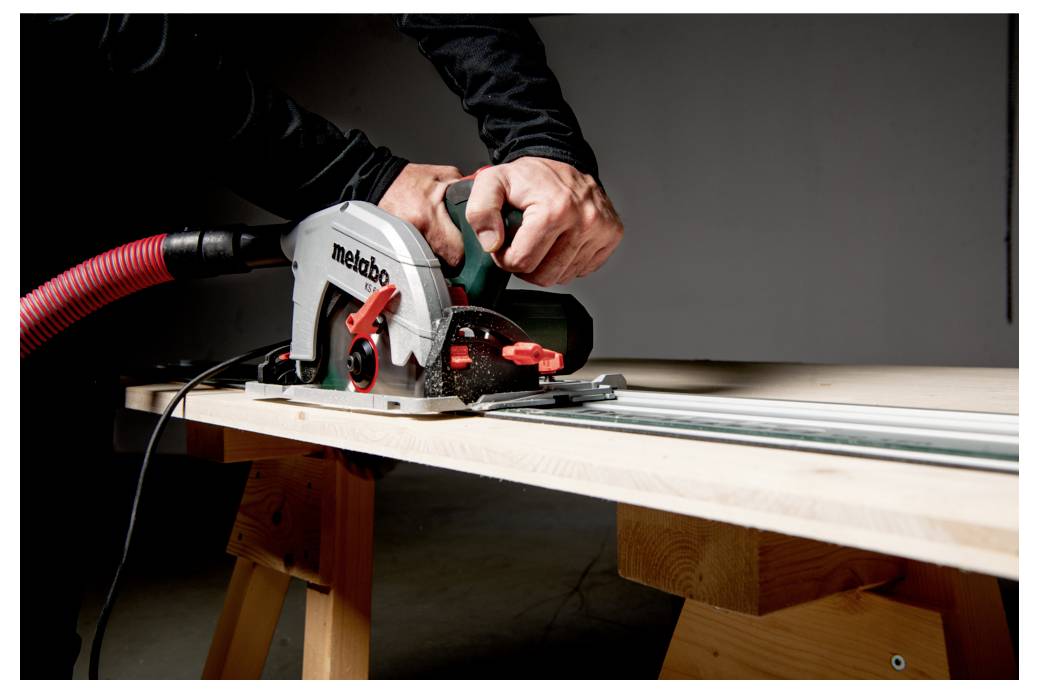 Eine Person, die eine Metabo-Kreissäge auf einem Holzbrett verwendet und sich auf präzises Sägen konzentriert. Die Säge ist mit einem roten Staubsaugeschlauch zur Staubsammlung verbunden.