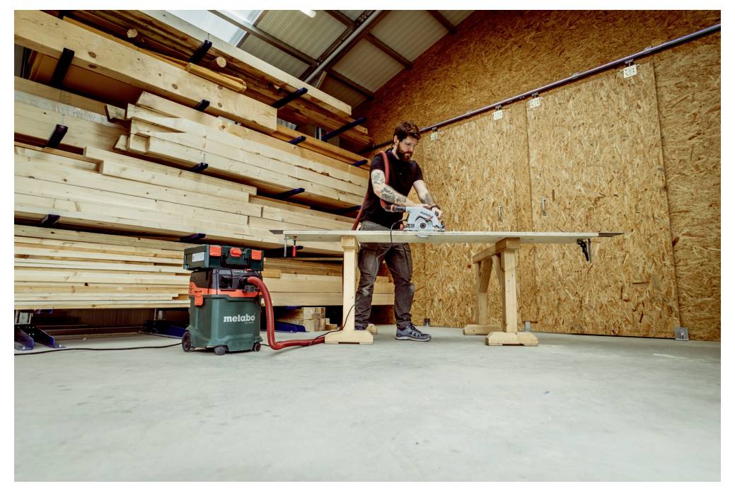 Eine Person verwendet eine Kreissäge, um Holz auf einer Werkbank in einer Werkstatt zu sägen, mit Holzstapeln im Hintergrund.