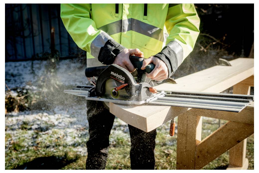 Eine Person, die eine Metabo-Kreissäge verwendet, um ein Holzbrett im Freien zu schneiden, trägt dabei eine leuchtend grüne Warnweste und Handschuhe.