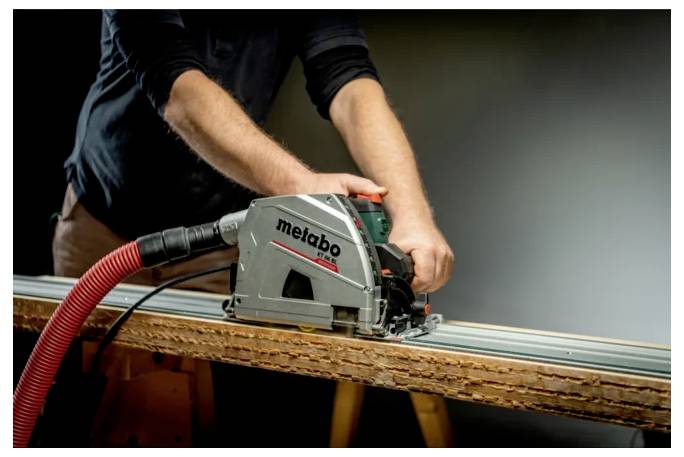 Person, die eine Metabo-Kreissäge mit einem Staubsaugerschlauch verwendet, um ein Holzbrett auf einer Werkbank zu sägen.