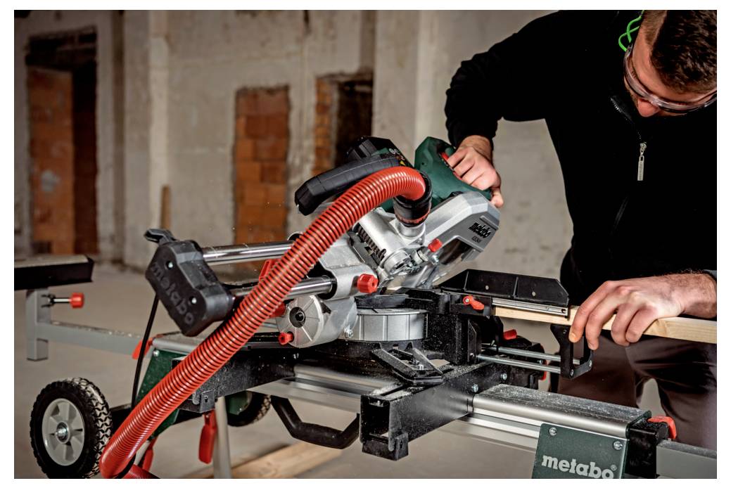 Eine Person bedient eine Kapp- und Gehrungssäge auf einer Baustelle und schneidet ein Holzstück. Die Umgebung ist im Innenbereich mit unfertig gemauerten Ziegelwänden.