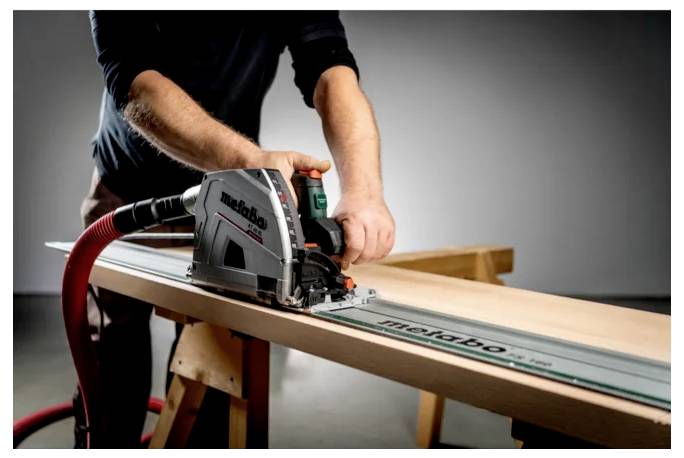 Eine Person benutzt eine Kreissäge auf einem langen Holzbrett und schneidet es entlang einer Führungsschiene in einer Werkstatt.