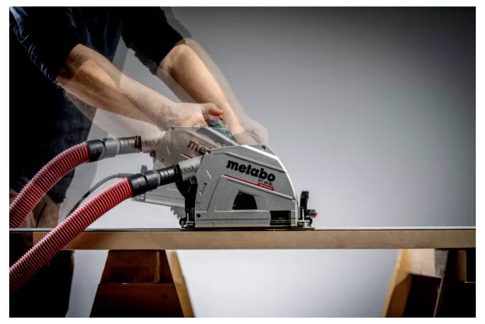 Eine Person, die ein Metabo-Elektrowerkzeug mit roten Absaugschläuchen verwendet, um entlang der Kante eines Holzbretts auf einer Werkbank zu schneiden oder zu schleifen.