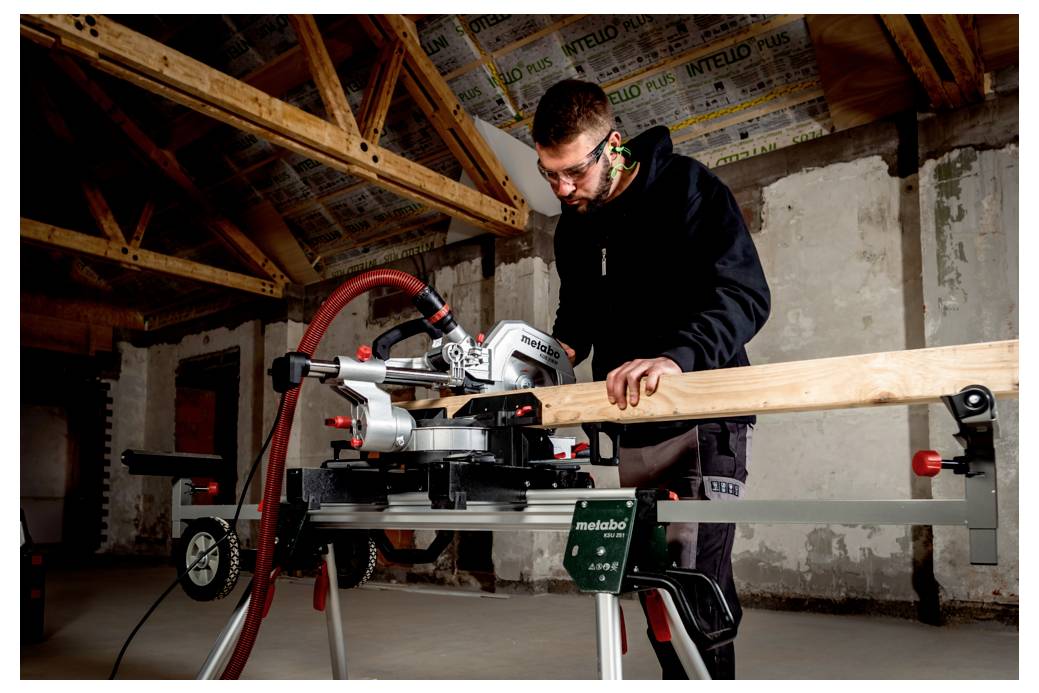 Eine Person, die eine Gehrungssäge verwendet, um einen Holzbalken in einem teilweise fertiggestellten Raum mit sichtbaren Balken und Dämmung an der Decke zu schneiden.