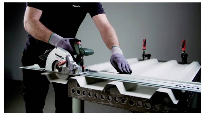 Eine Person benutzt eine Kreissäge, um ein Metallblech auf einer Werkbank zu schneiden, trägt Handschuhe und hält es mit Spannzwingen fest.