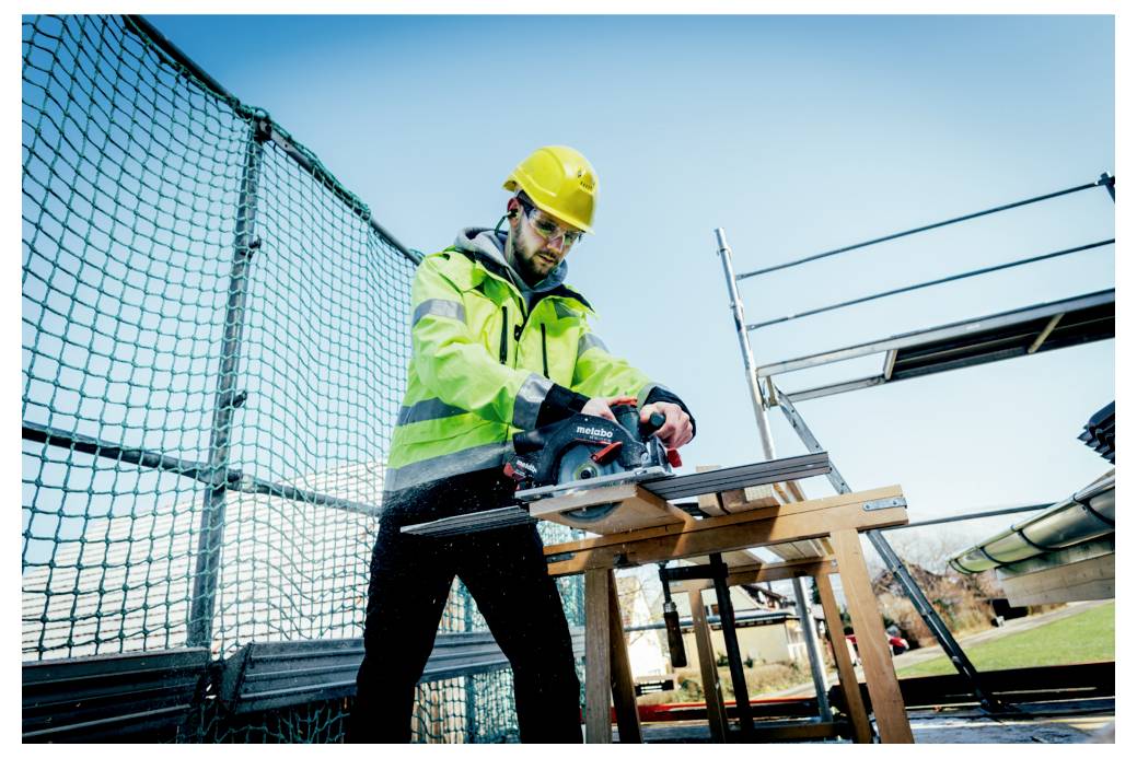Ein Bauarbeiter in Schutzausrüstung sägt mit einer Elektrosäge Holzbretter auf einer Baustelle, wobei im Hintergrund Gerüste und ein Zaun zu sehen sind.