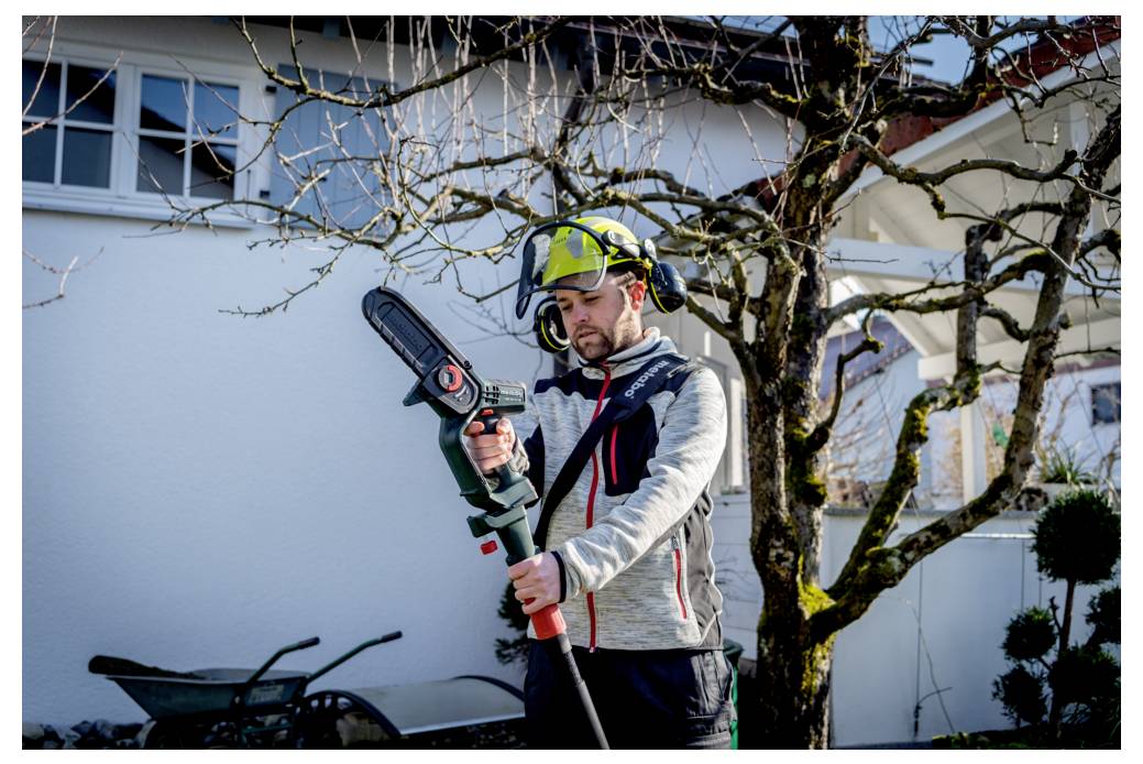 Eine Person mit Sicherheitsausrüstung bedient eine Teleskop-Säge, um Äste in einem Garten zu beschneiden, wobei ein weißes Haus und eine Schubkarre im Hintergrund zu sehen sind.