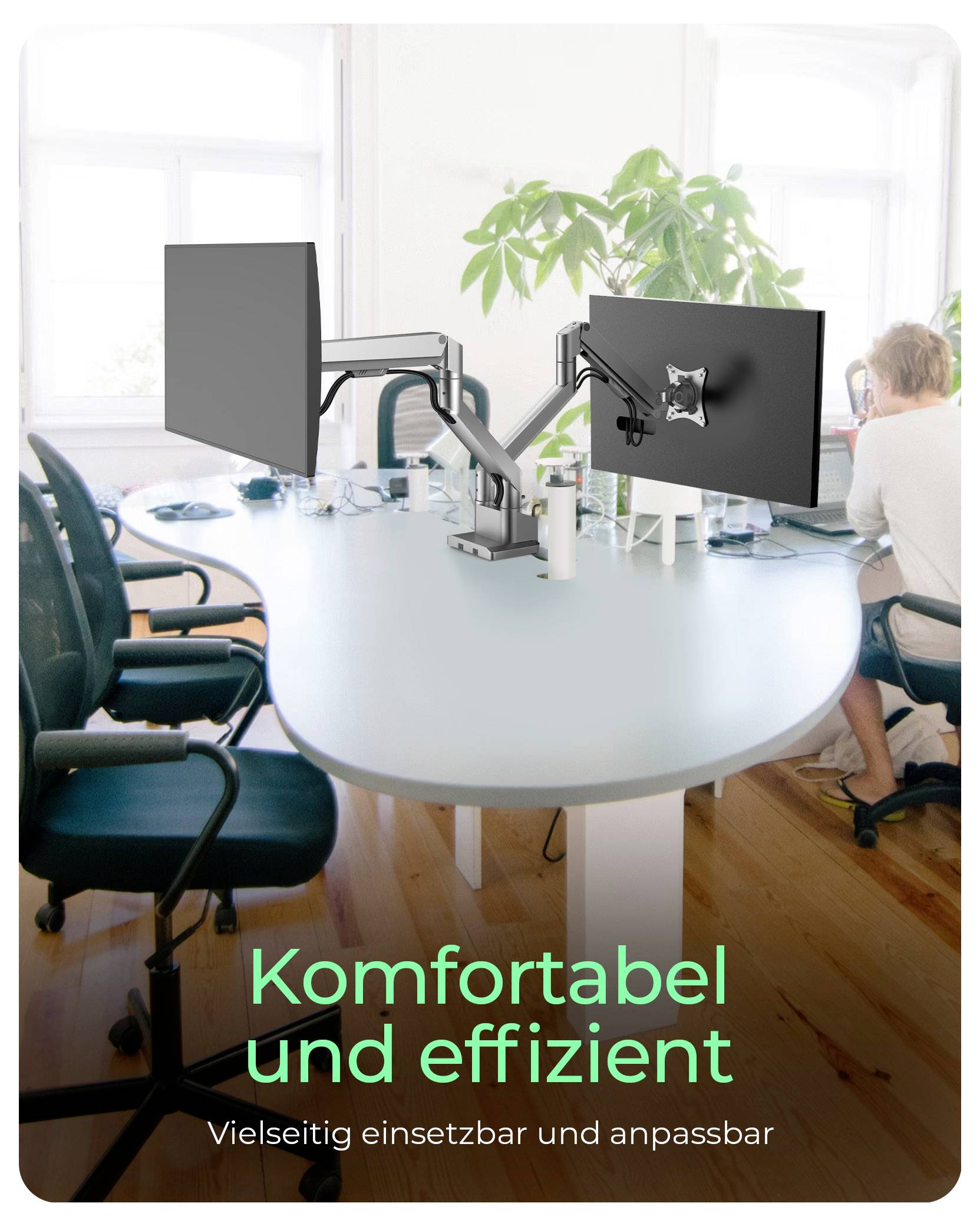 Zwei Computerbildschirme an einem ergonomischen Arm über einem runden Bürotisch. Im Hintergrund tippt eine Person auf einem Laptop.