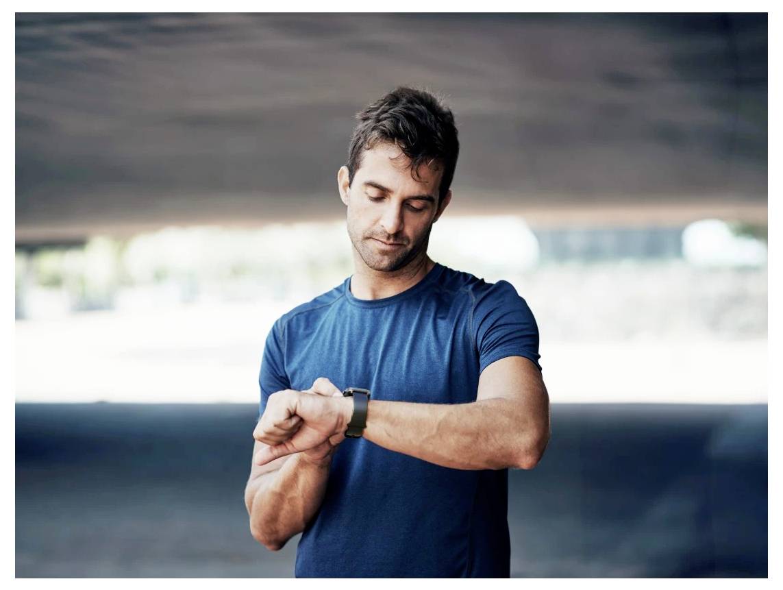 Ein Mann in einem blauen Hemd schaut auf seine Armbanduhr im Freien, was darauf hindeutet, dass er möglicherweise die Zeit verfolgt oder seine Aktivität überwacht.