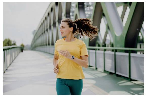 Eine Frau joggt über eine Brücke mit grünen Geländern, trägt ein gelbes Shirt und petrolblaue Leggings, ihr Haar weht im Wind.