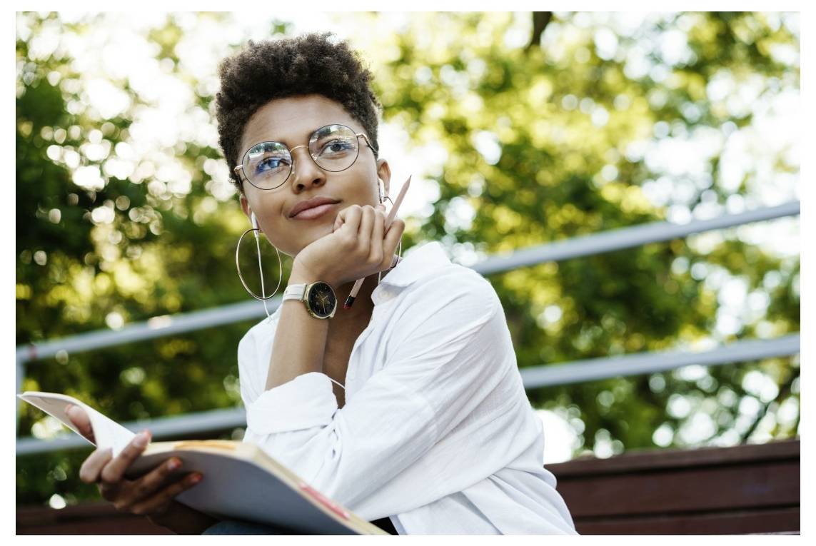 Eine Person sitzt im Freien, trägt eine Brille und hält ein Notizbuch und einen Stift, und wirkt nachdenklich, während Bäume im unscharfen Hintergrund zu sehen sind.