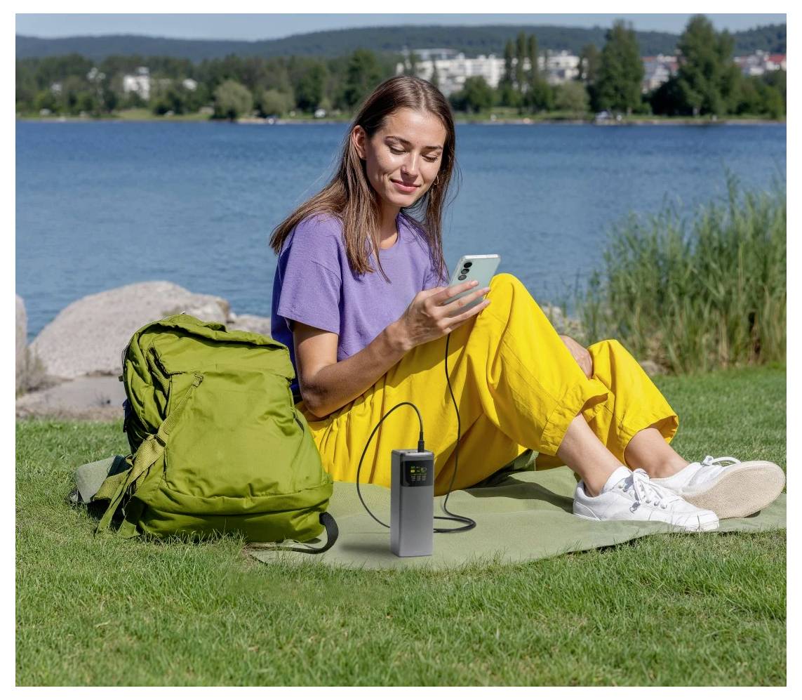 Eine Frau sitzt auf Gras am See und benutzt ein Smartphone, das an einem tragbaren Solarladegerät angeschlossen ist. Neben ihr liegt ein grüner Rucksack.