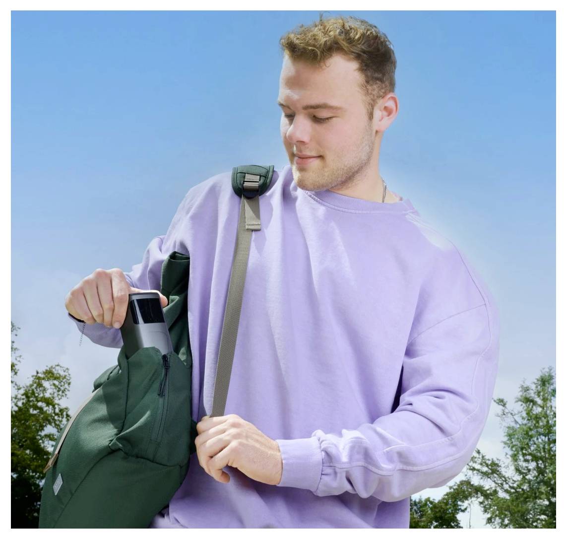 Eine Person in einem hellvioletten Sweatshirt legt eine Wasserflasche in einen grünen Rucksack im Freien, mit einem klaren blauen Himmel und Bäumen im Hintergrund.
