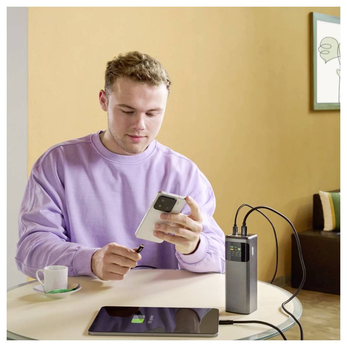 Eine Person sitzt an einem Tisch und verbindet ein Telefon mit einer Powerbank, während ein weiteres Telefon neben einer Kaffeetasse lädt. Im Hintergrund befindet sich ein gemütlicher Raum.