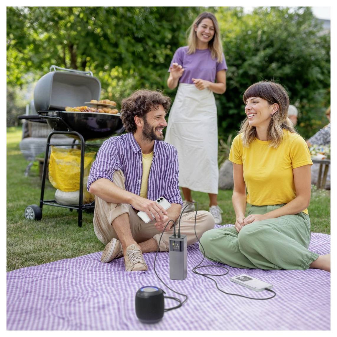 Ein Mann und eine Frau sitzen auf einer Picknickdecke, lächeln und unterhalten sich. Ein Barbecue-Grill befindet sich im Hintergrund, und eine weitere Frau steht daneben.