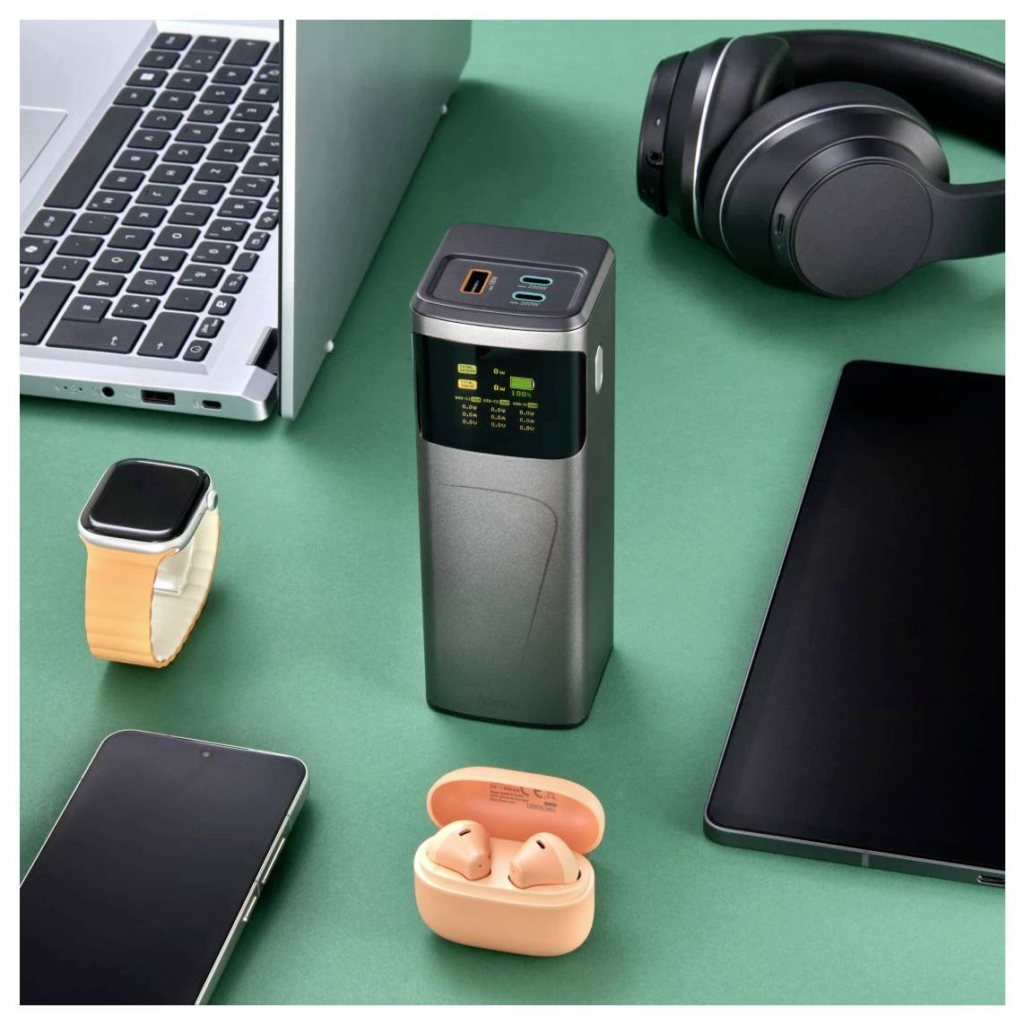 Desktop-Setup mit einem geschlossenen Laptop, Kopfhörern, Smartwatch, Smartphone, Tablet, orangefarbenen Ohrhörern und einem tragbaren Ladegerät auf einer grünen Oberfläche.
