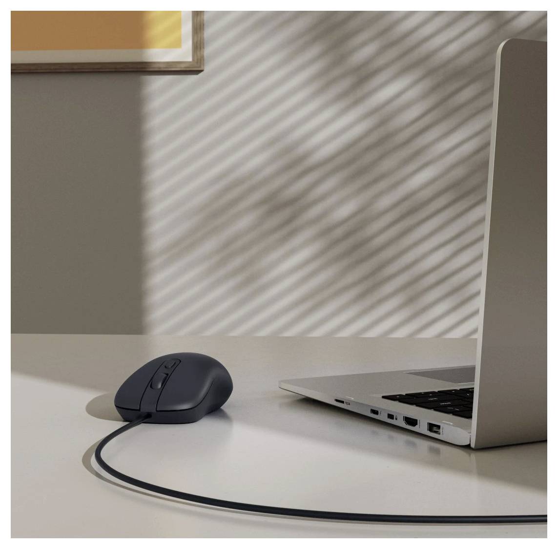 Eine schlanke schwarze Computermaus, die mit einem silbernen Laptop auf einem weißen Schreibtisch verbunden ist, während Sonnenlicht gestreifte Schatten durch ein Fensterrollo wirft.