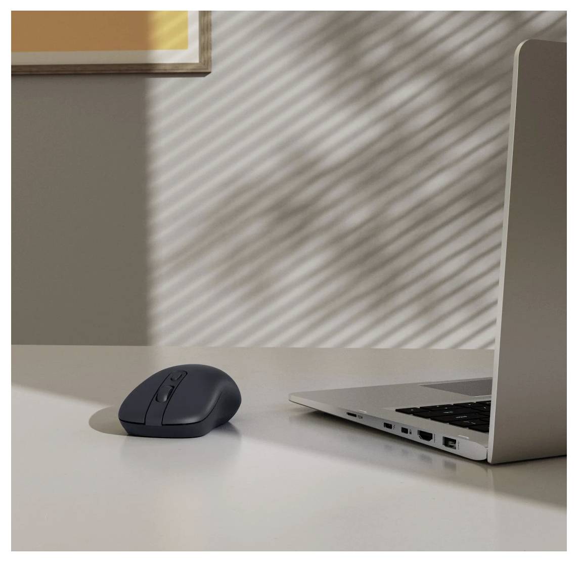 Eine Computermaus neben einem geöffneten silbernen Laptop auf einem weißen Tisch; Sonnenlicht erzeugt gestreifte Schatten an der Wand im Hintergrund.