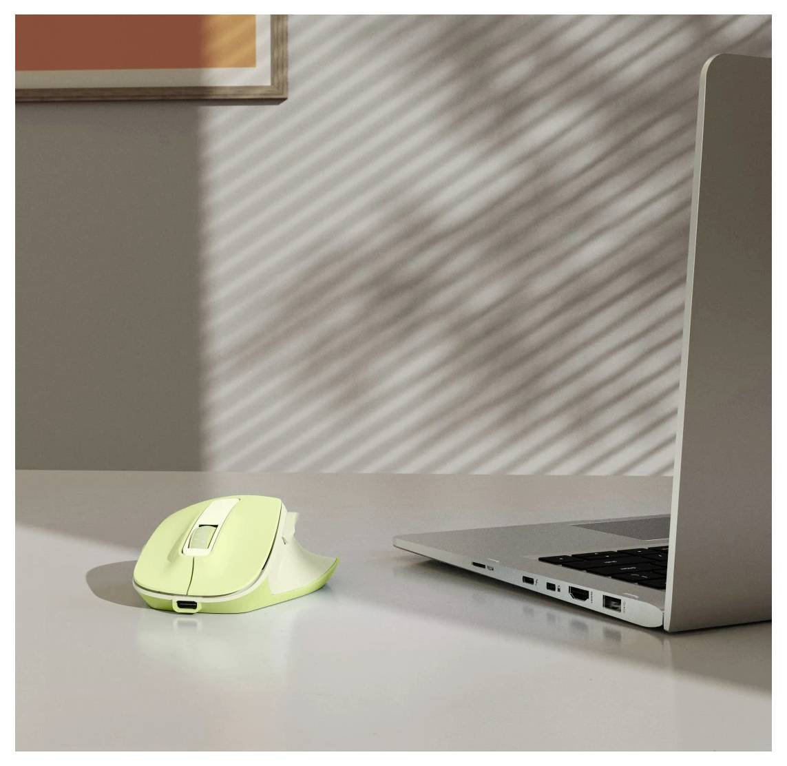 Eine grüne kabellose Computermaus, die neben einem geöffneten Laptop auf einem Tisch liegt, während Sonnenlicht gestreifte Schatten durch Jalousien wirft.