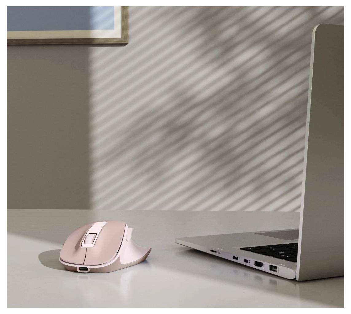 Eine pinke ergonomische drahtlose Maus auf einem weißen Schreibtisch neben einem geöffneten silbernen Laptop, während Sonnenlicht gestreifte Schatten durch ein Fenster wirft.