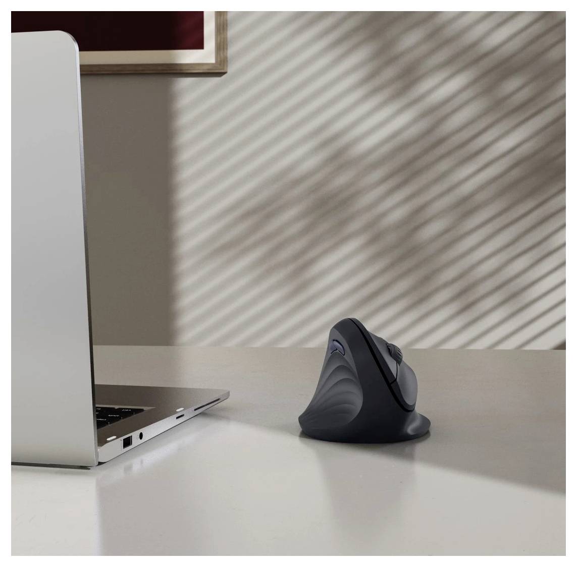 Ein Laptop und eine ergonomische Maus befinden sich auf einem weißen Tisch und werden von weichem, gestreiftem Licht beleuchtet, das durch Fensterjalousien fällt.