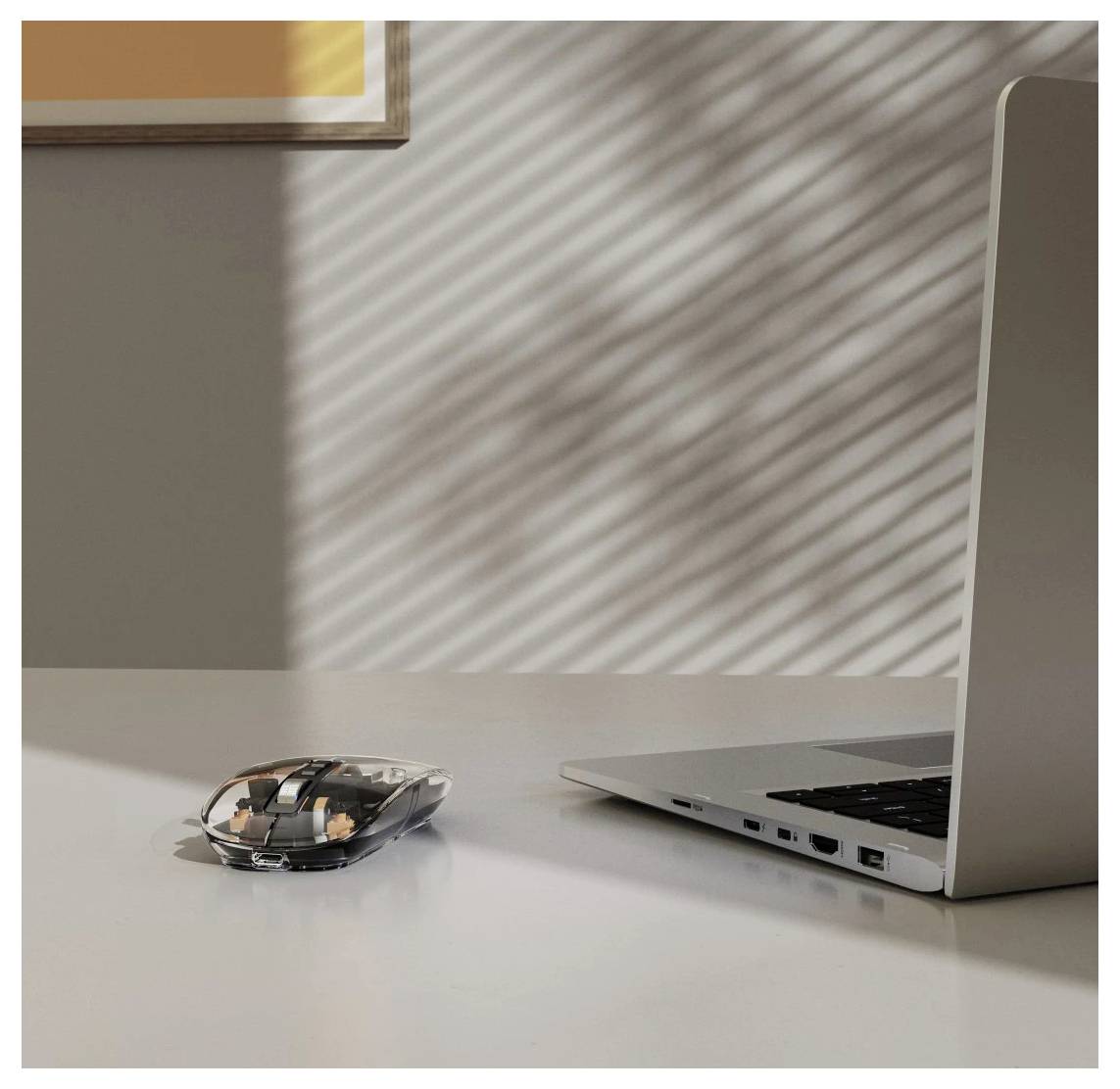 Eine schlanke, transparente Computermaus neben einem silbernen Laptop auf einem sonnendurchfluteten Schreibtisch, mit Schattenmustern von Jalousien an der Wand.