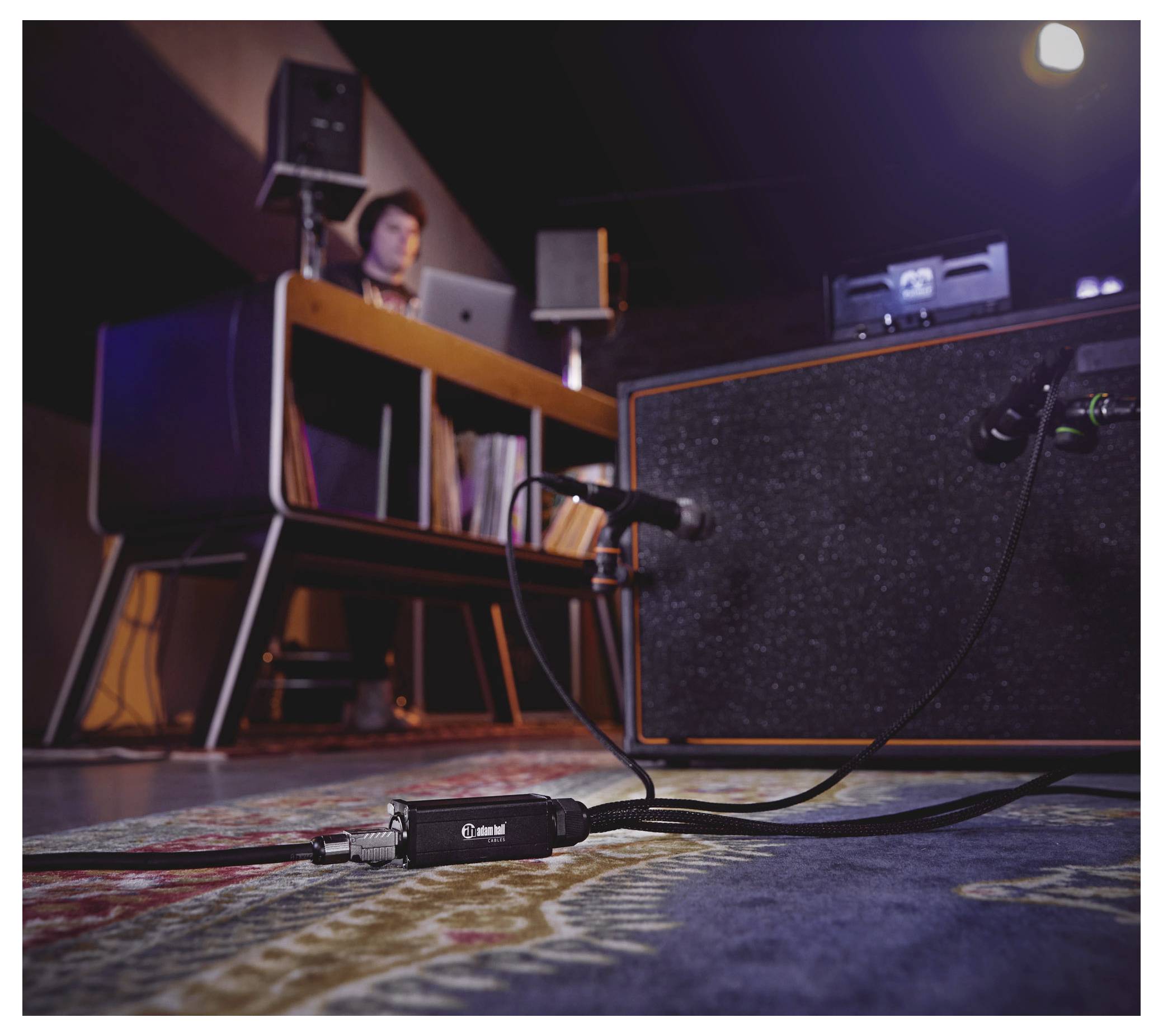 Ein Aufnahmestudio mit Fokus auf einem Gitarrenpedal und Kabel auf einem Teppich, verbunden mit Ausrüstung. Der Hintergrund zeigt eine Person an einem Schreibtisch mit Audioausrüstung.