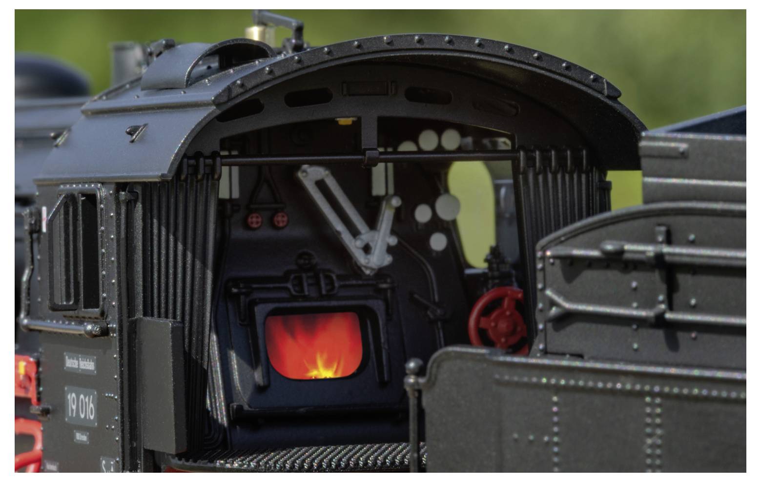 Das Bild zeigt das Innere der Führerkabine einer Dampflokomotive mit einem hell lodernden Feuer im Feuerbüchsenraum und detaillierten mechanischen Bedienungselementen.