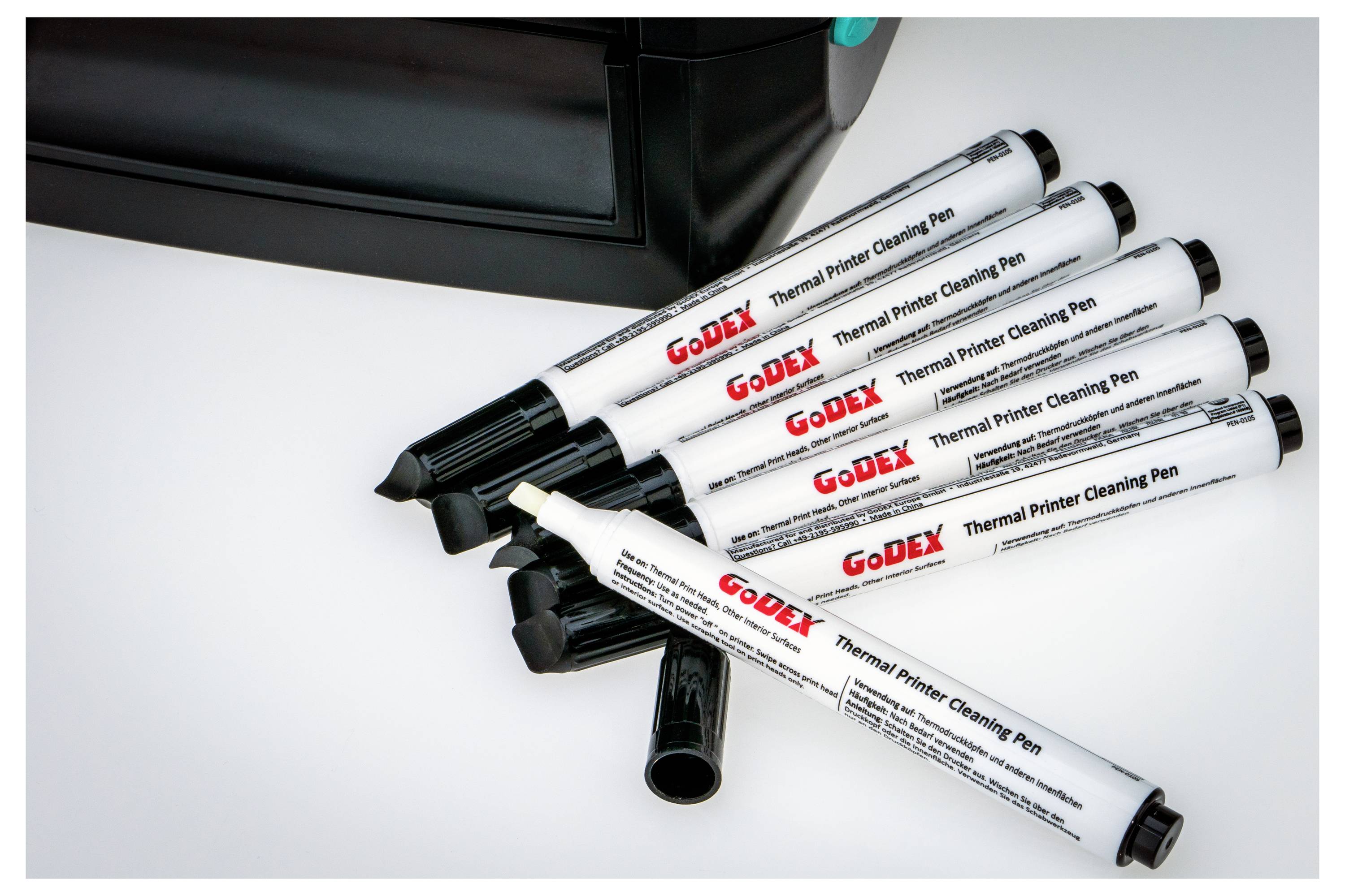 Mehrere thermische Druckerreinigungsstifte sind auf einer weißen Oberfläche neben einem schwarzen Drucker verstreut und zeigen ihre Verwendung bei der Druckerwartung.