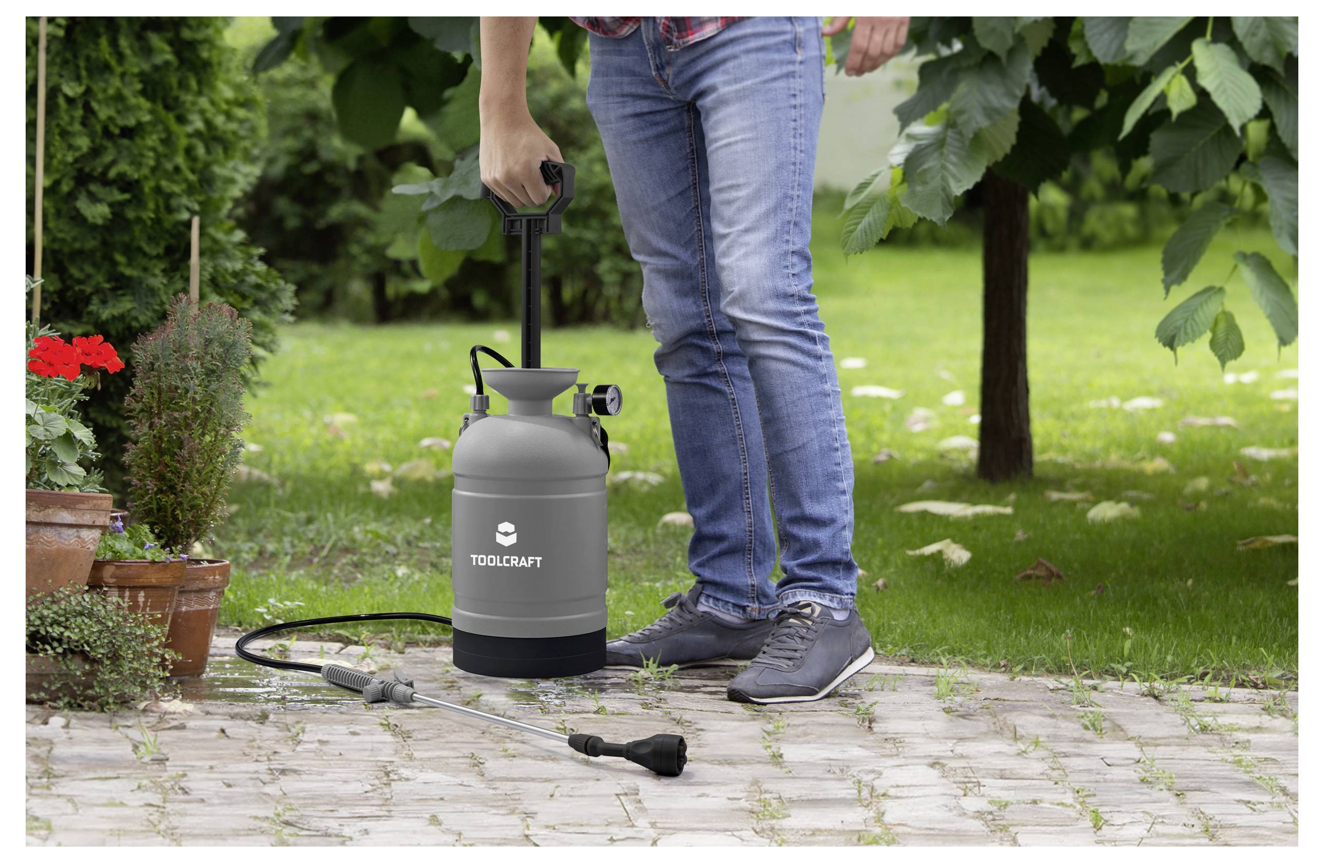 Eine Person, die eine Handspritzpumpe in einem Garten verwendet, umgeben von Töpfen und grünen Pflanzen. Die Spritzpumpe trägt das Etikett „Toolcraft