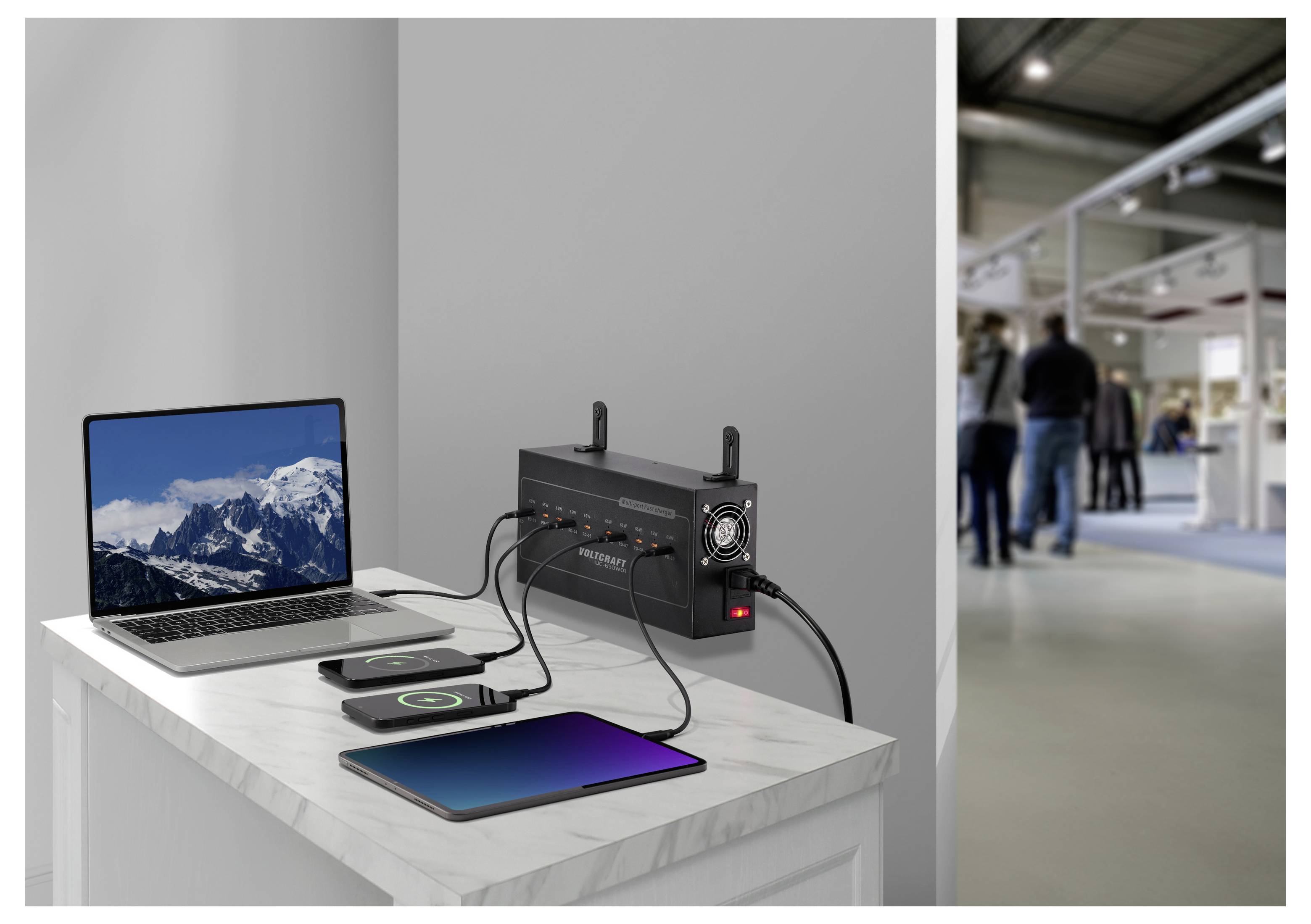 Ein Zimmer mit Geräten, die auf einem Schreibtisch aufgeladen werden: Ein Laptop, Tablet und Smartphone sind an einer an der Wand angebrachten Ladestation angeschlossen.