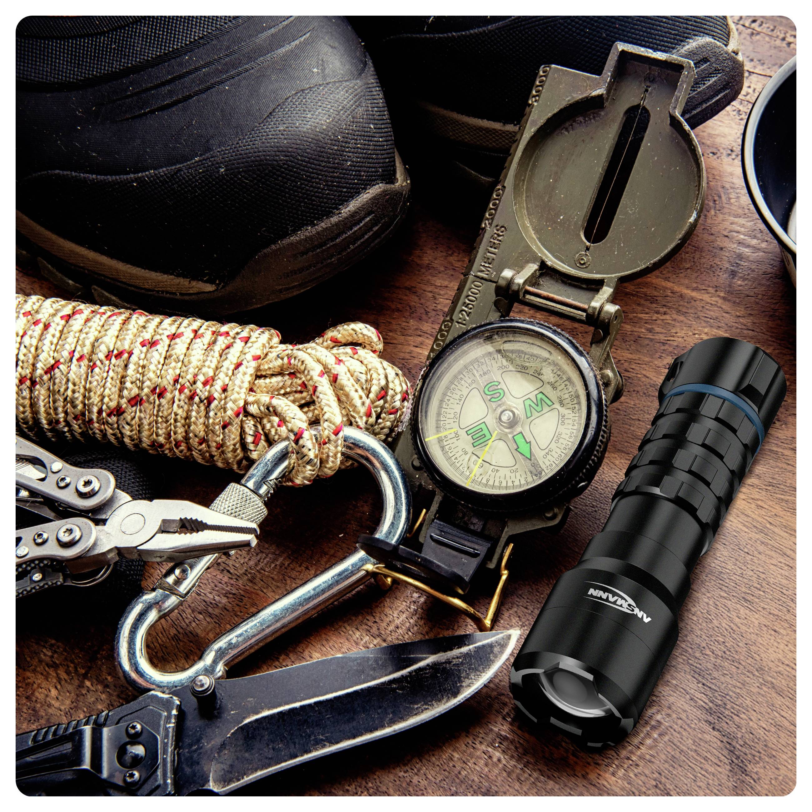 Outdoor-Ausrüstung einschließlich eines Wanderschuhs, Kompasses, Taschenlampe, Multitools, Karabiners, Seils und Messers auf einer Holzoberfläche, was Abenteuerlust andeutet.