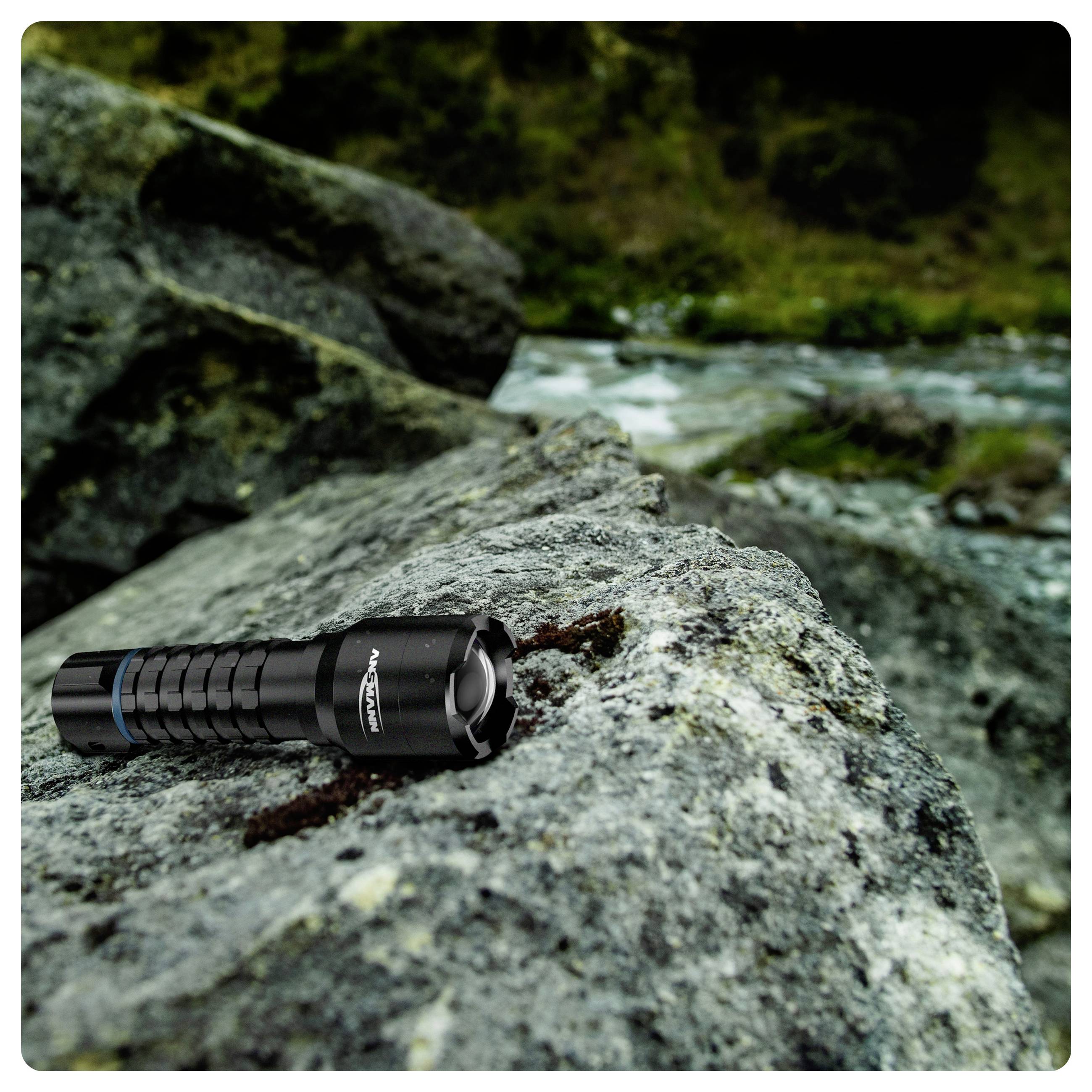 Eine schwarze Taschenlampe liegt auf einem großen, grauen Felsen in einer üppigen Außenumgebung in der Nähe eines fließenden Flusses.