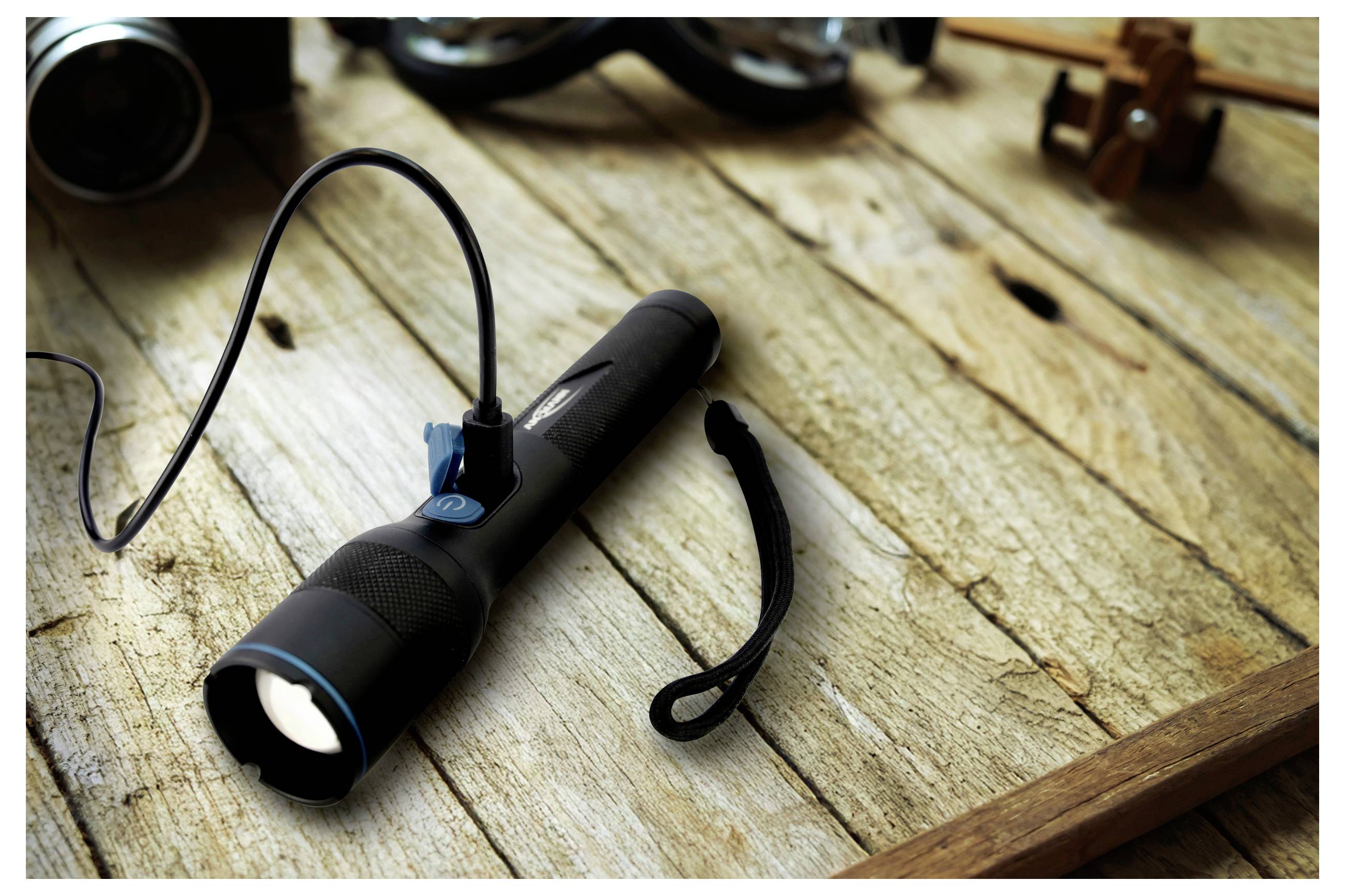 Eine schwarze Taschenlampe, die mit einem Ladekabel verbunden ist, liegt auf einer rustikalen Holzoberfläche neben kleinen Gegenständen und vermittelt eine Vintage-Atmosphäre.