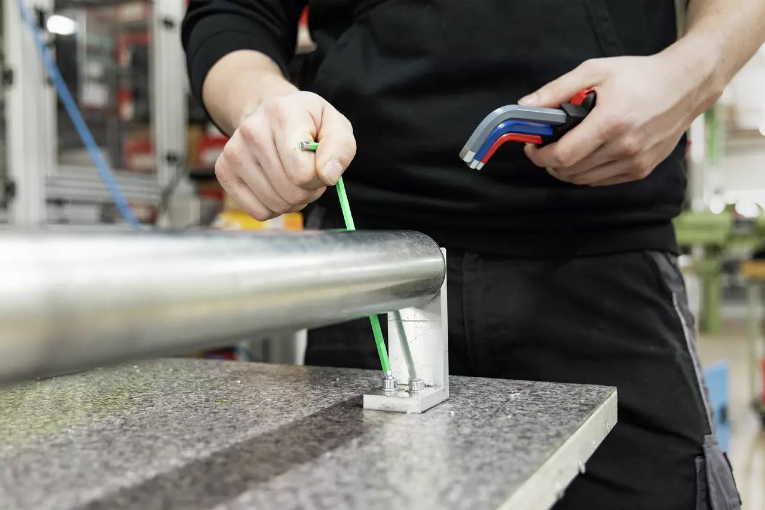 Eine Person zieht Schrauben an einer zylindrischen Metallstange mit einem Sechskantschlüssel, wobei sie technische Arbeit in einem industriellen Umfeld demonstriert.