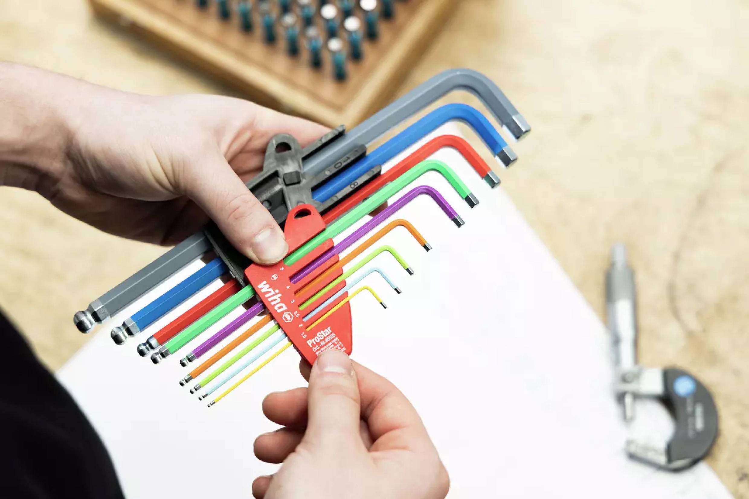 Hände, die einen Satz bunter Sechskantschlüssel in verschiedenen Größen halten, angeordnet auf einem fächerartigen Halter. Ein Mikrometer ist im Hintergrund sichtbar.