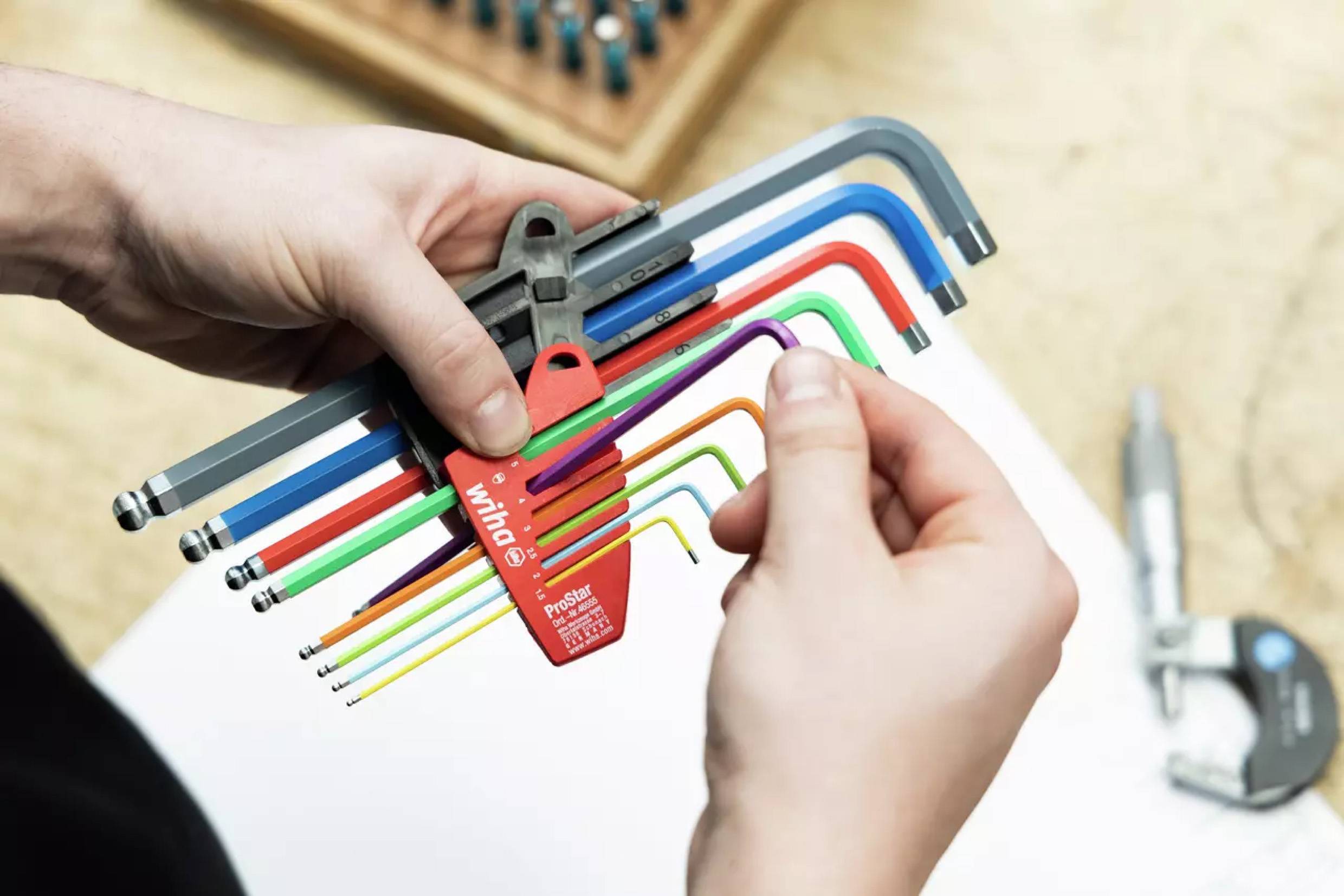 Eine Person hält einen Satz bunter Sechskantschlüssel in Rot, Grün, Blau und Orange vor einem weiß-beigen Hintergrund, was eine Werkzeugauswahl andeutet.