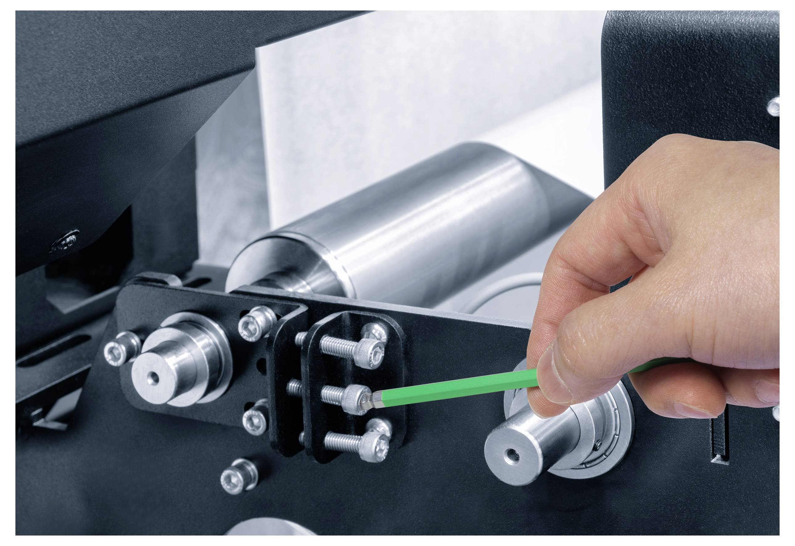 Eine Hand verwendet ein grünes Werkzeug, um Bolzen an einer Industriemaschine einzustellen, wobei der Fokus auf Wartungs- oder Montageaufgaben liegt und Präzisionsarbeit betont wird.